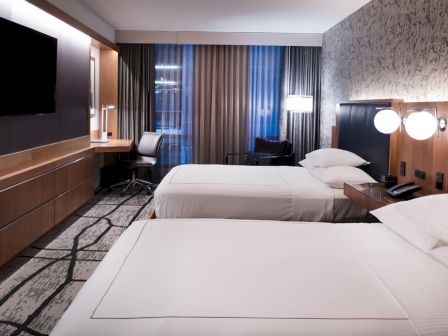 A modern hotel room with two beds, a TV, a desk with a chair, and stylish lighting and decor on the walls.