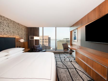 A modern hotel room featuring a large bed, desk, chair, and a window with a city view. A TV is mounted on a wooden accent wall.