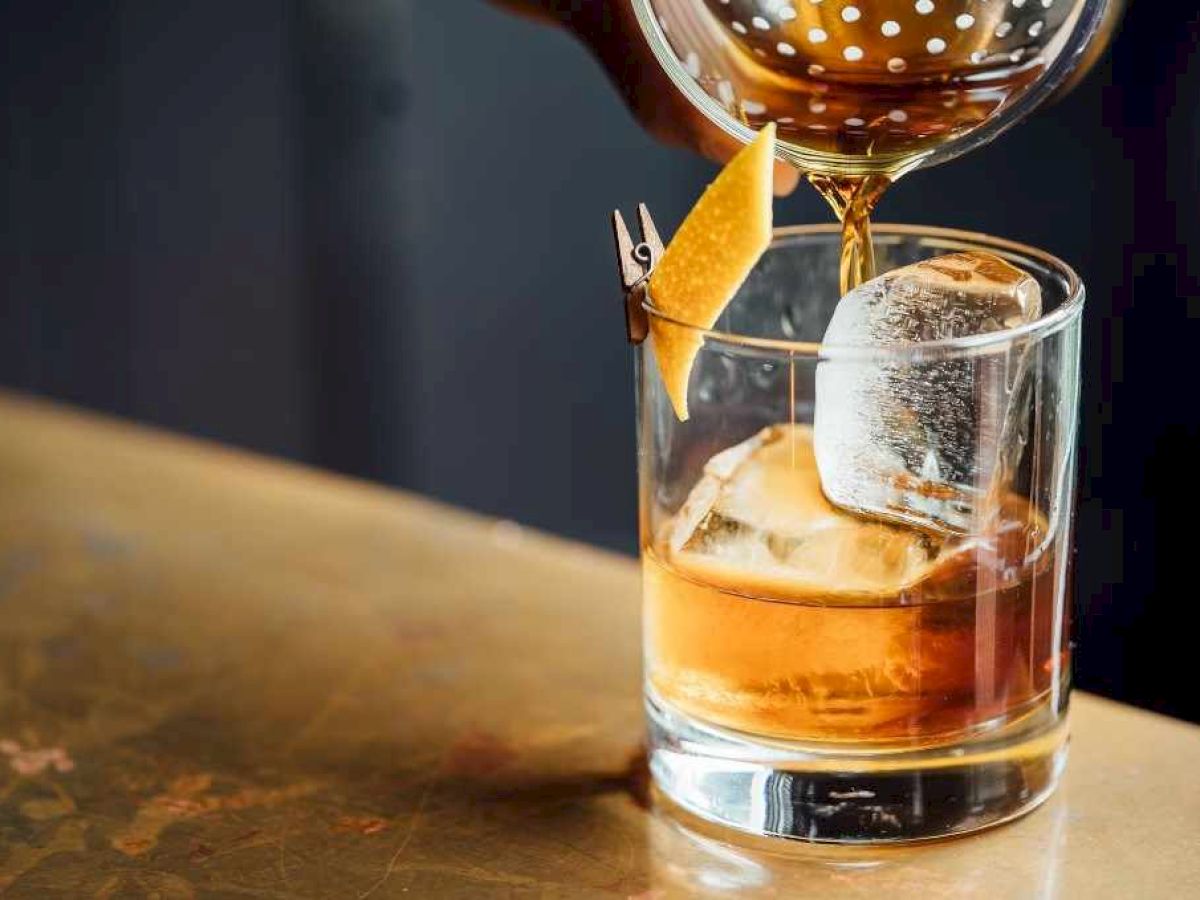 A glass with a brown cocktail, garnished with a lemon peel and large ice cube, being poured from a strainer.