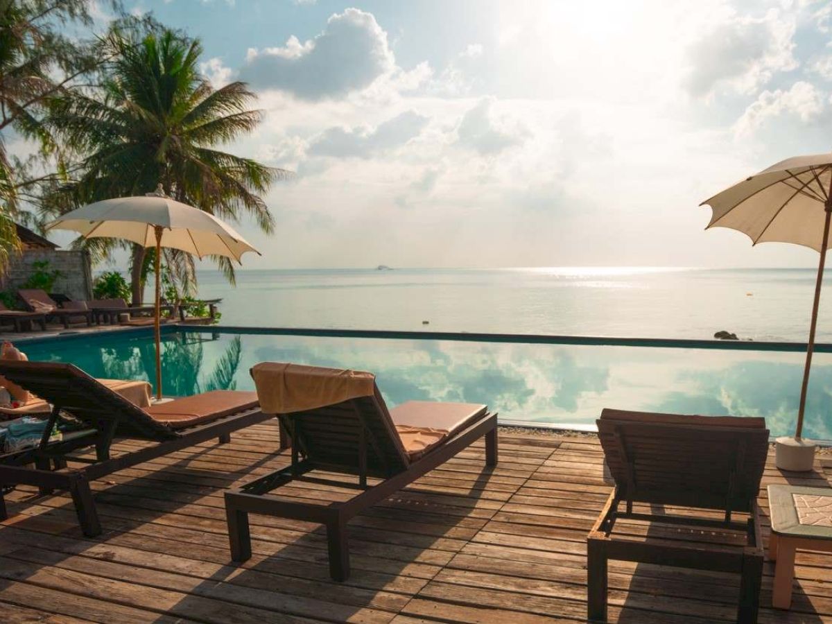 Lounge chairs and umbrellas by a pool overlook a serene sea view under a partly cloudy sky with palm trees in the background.