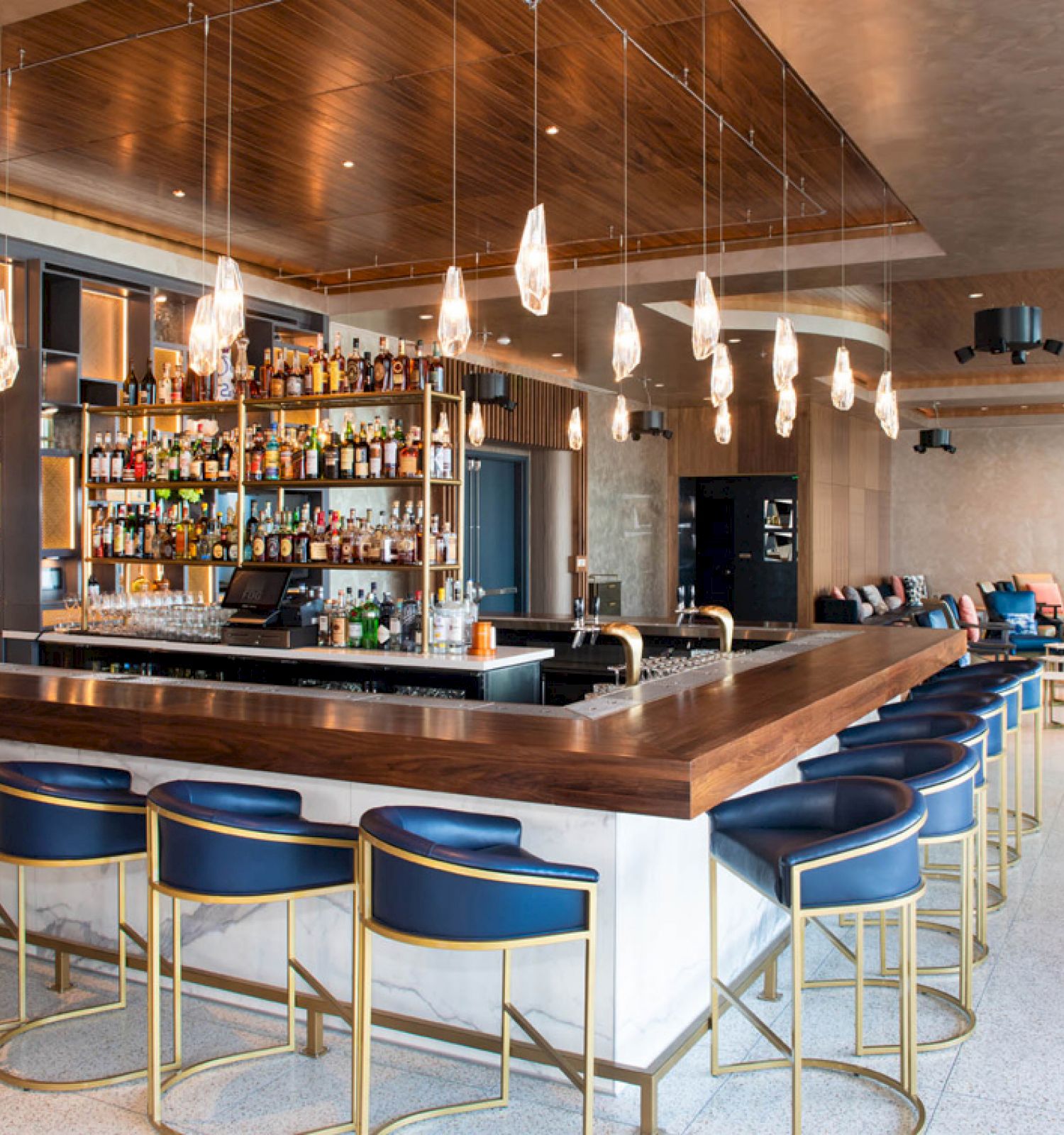 A modern bar with a wooden counter, high chairs, pendant lights, and shelves stocked with bottles. It has a stylish lounge area in the back.