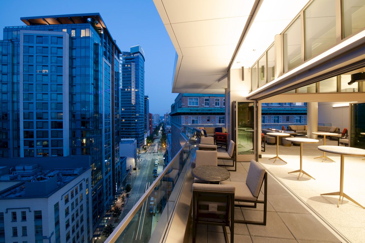 A modern balcony overlooks a city street at dusk, with tables and chairs arranged neatly along the glass railing and bright interior lights.