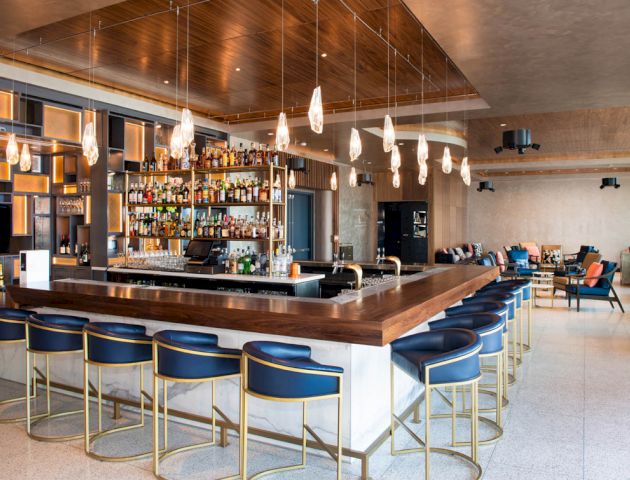 The image shows a modern bar with a large counter, blue chairs, hanging lights, and a wide selection of liquor bottles on shelves.