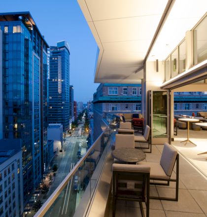 A modern rooftop patio with tables and chairs overlooks a cityscape of tall buildings, captured during evening.