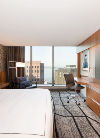 A modern hotel room with a bed, desk, chair, large TV, and a window view of buildings and the water.