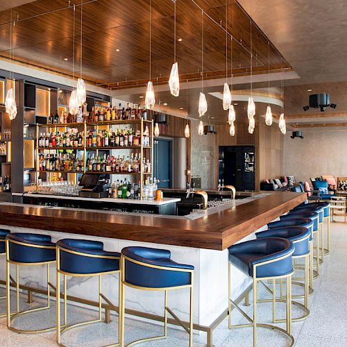A modern bar with a wooden counter, blue stools, and pendant lights, surrounded by shelves of bottles and a stylish, spacious interior.