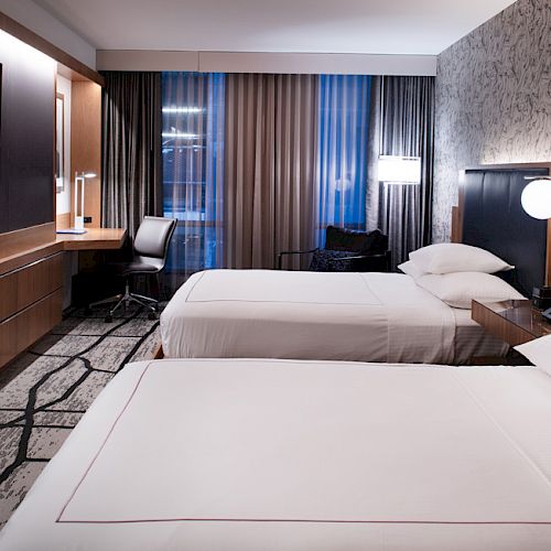 The image shows a modern hotel room with two neatly made beds, a TV, a desk by the window, and stylish wall decor.