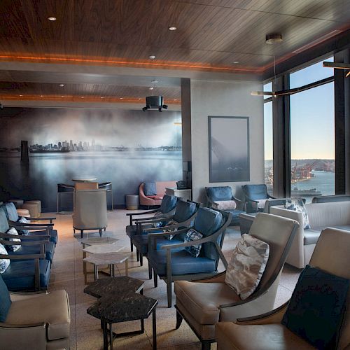 The image shows a luxurious lounge with plush seating, large windows offering a waterfront view, and modern decor.