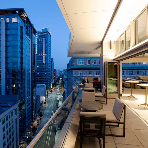 The image shows a modern cityscape with a balcony, tables, and chairs overlooking a street and tall buildings at dusk.