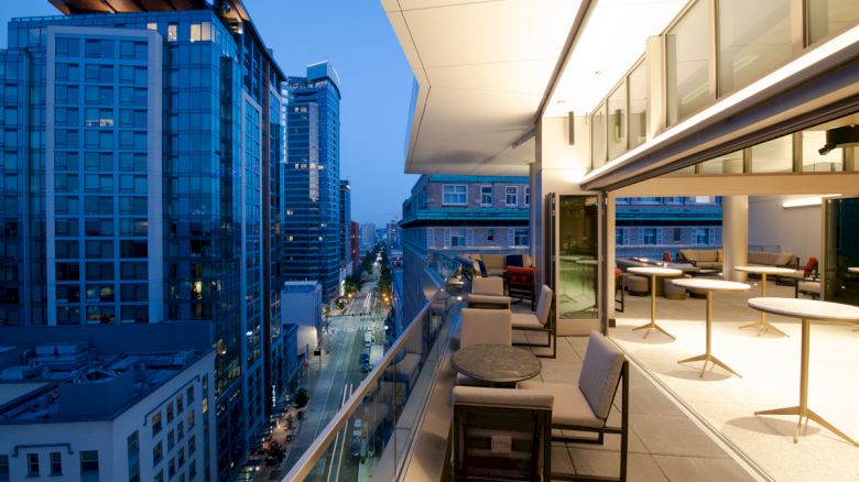 A modern rooftop terrace overlooking a cityscape at dusk, featuring seating and tables with surrounding tall buildings.