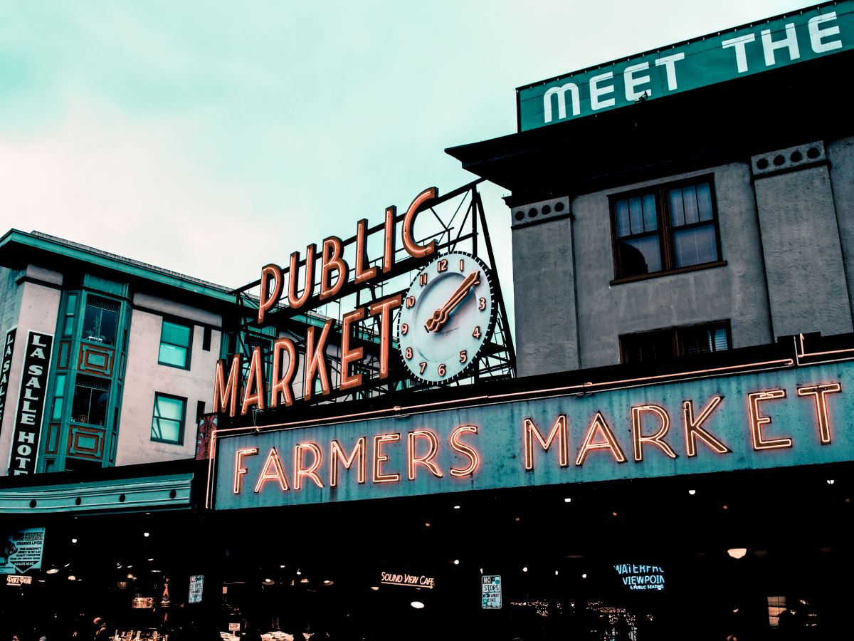 The image shows a public market with a neon sign reading "Farmers Market," featuring a clock, set against a building backdrop.