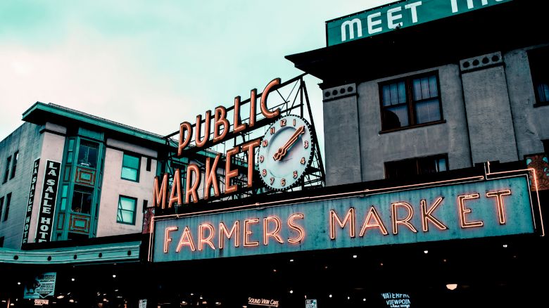 The image shows a public market with a neon sign reading "Farmers Market," featuring a clock, set against a building backdrop.