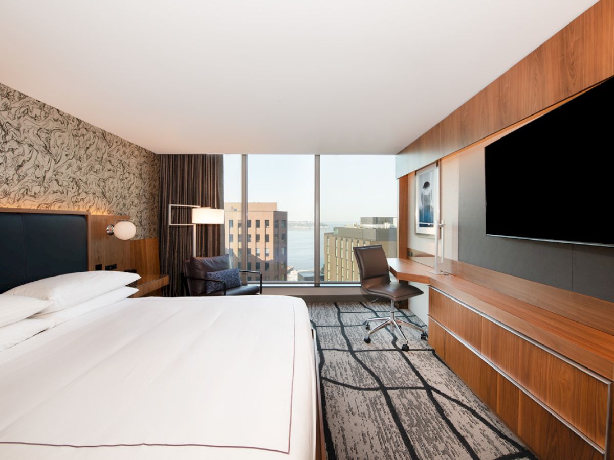 A modern hotel room features a large bed, desk, chair, wall-mounted TV, and a window with a city view.