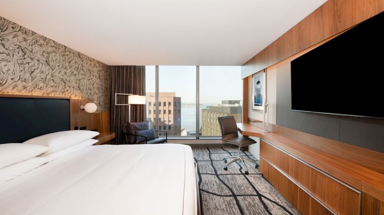 A modern hotel room features a large bed, desk, chair, wall-mounted TV, and a window with a city view.
