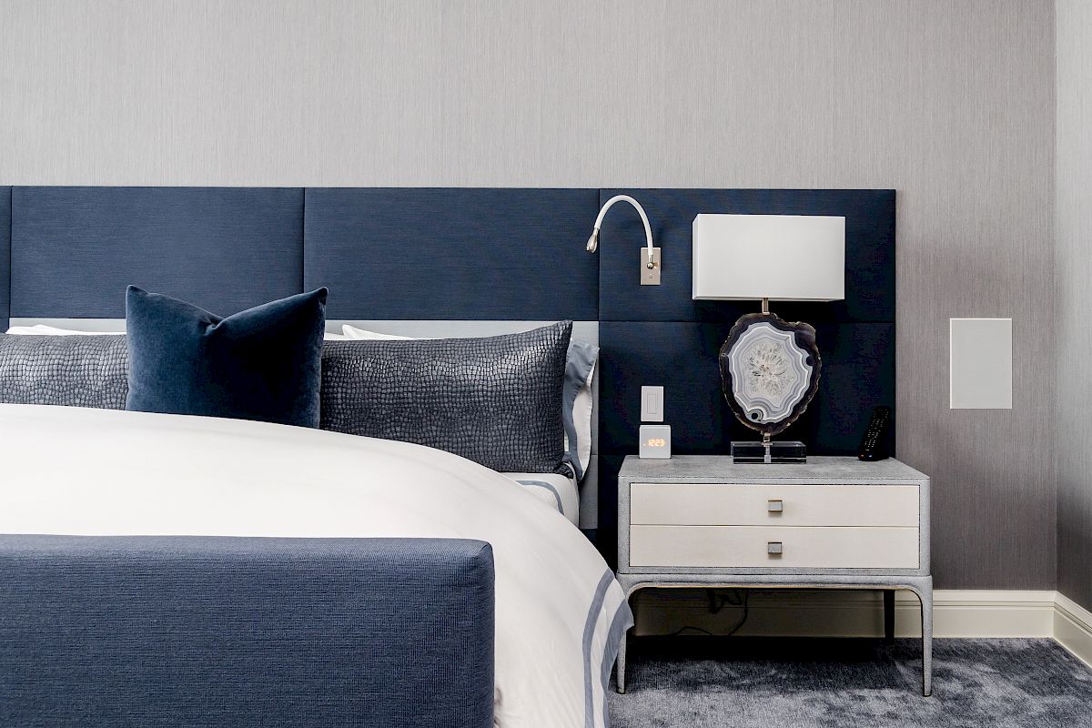 A modern bedroom with a blue and gray theme, featuring a bed, two pillows, a nightstand with a lamp, and a wall light.