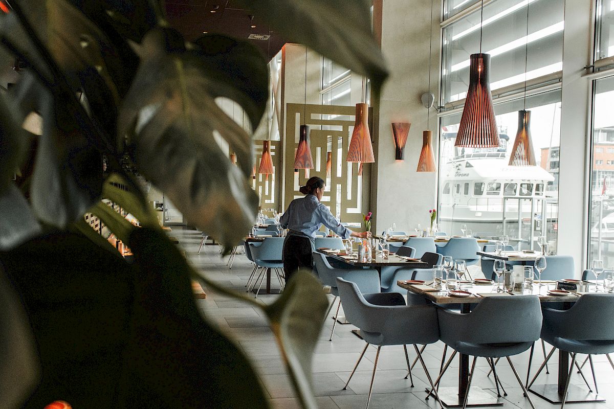 A restaurant interior with modern decor, a person setting tables, and large windows letting in natural light, captured through plant leaves.