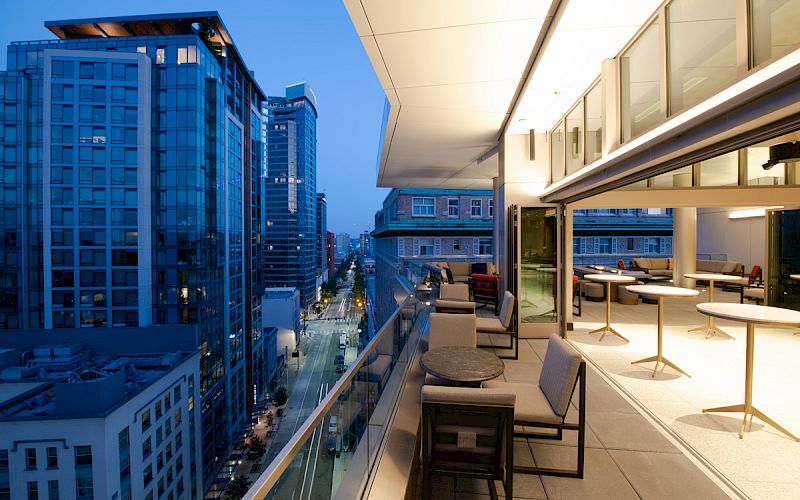 A modern rooftop patio with seating overlooks a cityscape of tall buildings at dusk, creating a stylish urban atmosphere.