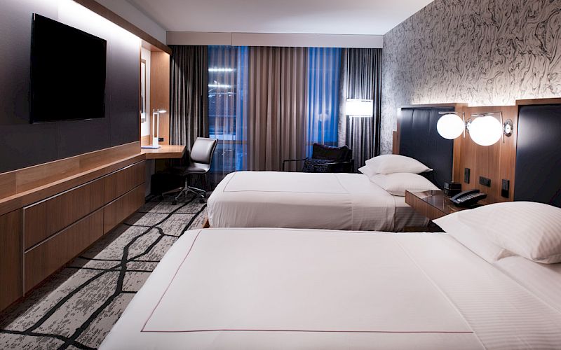 The image shows a modern hotel room with two neatly made beds, a desk, a TV, and stylish lighting and decor.