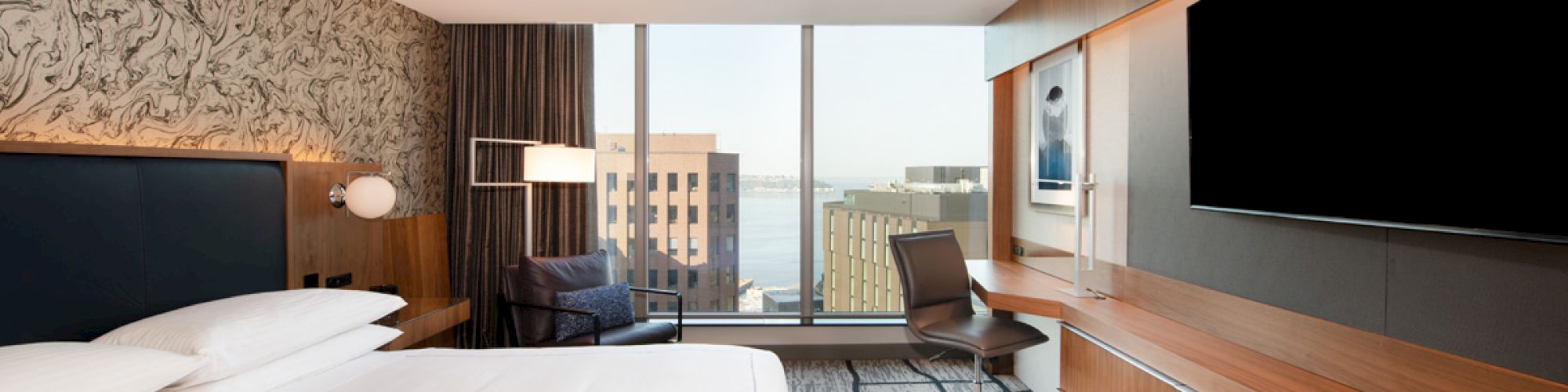 A modern hotel room with a large bed, wall TV, desk, chair, and city view through a window.